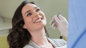 The image shows a woman smiling while sitting in a dental chair, with a dentist holding a dental mirror near her mouth. She appears happy and relaxed during her dental examination.