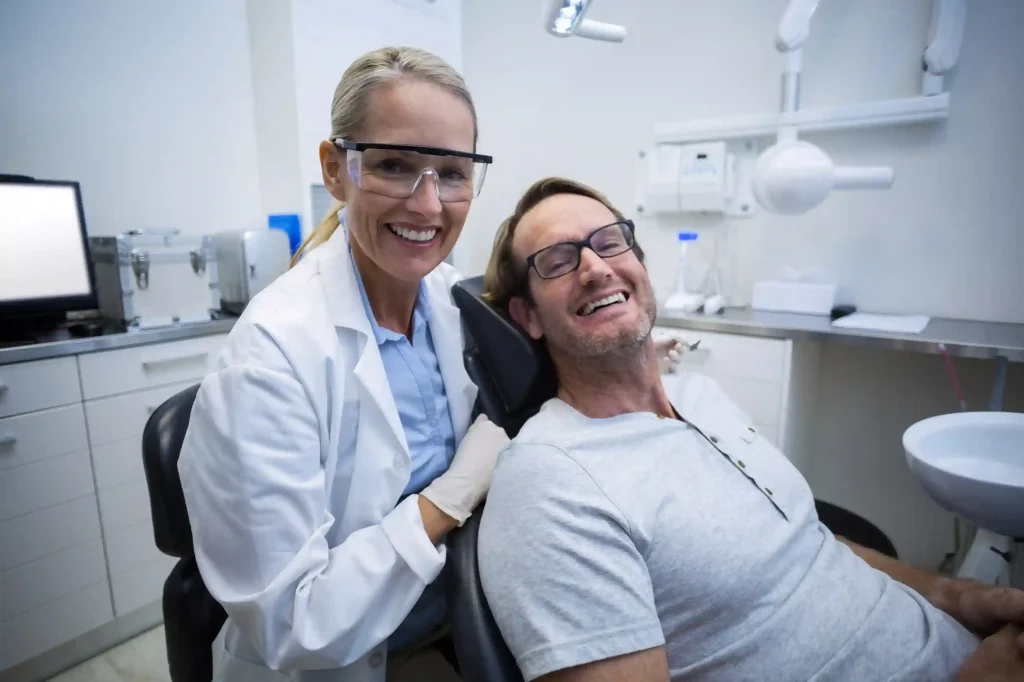 Dentist wearing protective eyewear smiling alongside a relaxed patient in a modern dental clinic. Represents a successful procedure of having a wisdom tooth taken out by an emergency dentist in London. - Liberty Dentists